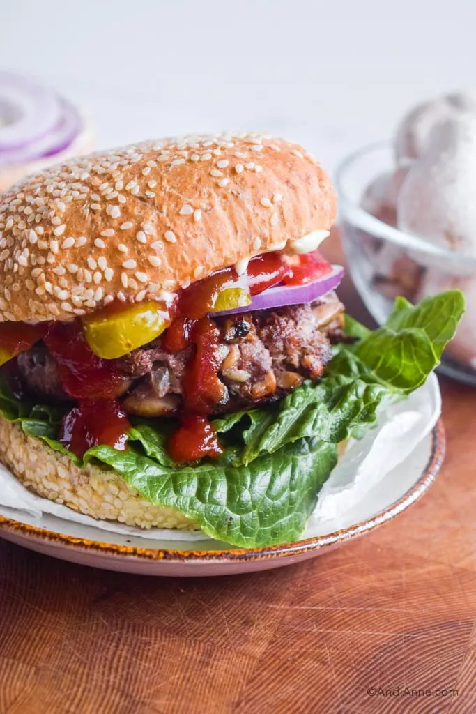 Burger with lettuce, ketchup, onion, pickle and mushroom beef burger. Bowl of white mushrooms in background.