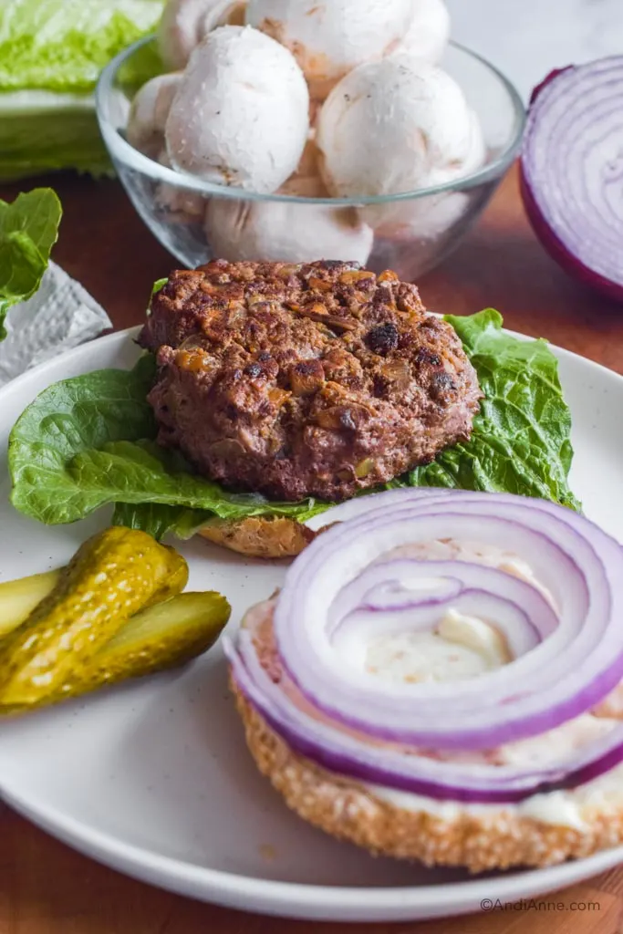 Burger patty sitting on lettuce and a bun with sliced onion and pickles in the front. 