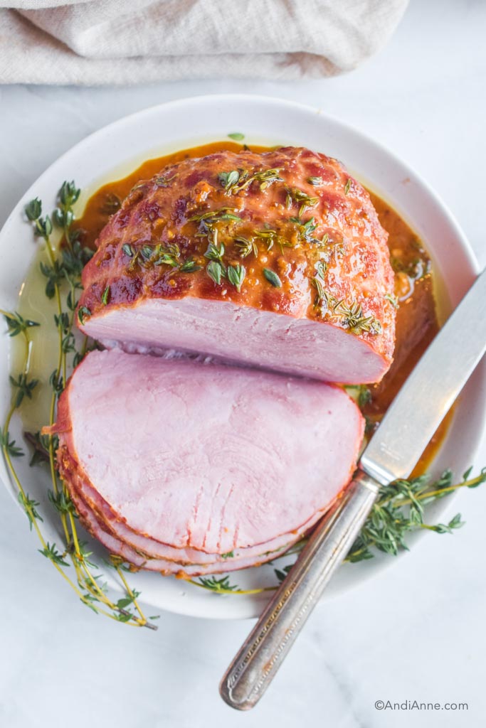 Looking down at honey mustard thyme glazed ham on a white plate with fresh thyme sprigs and a knife.