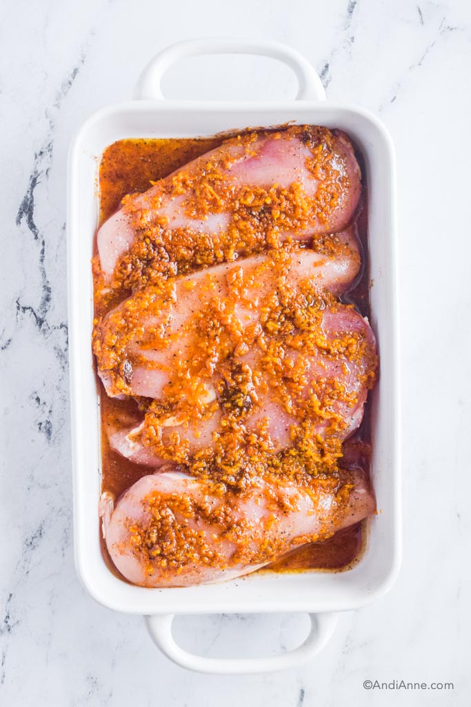 White casserole dish with four raw chicken breasts and orange sauce drizzled over top.
