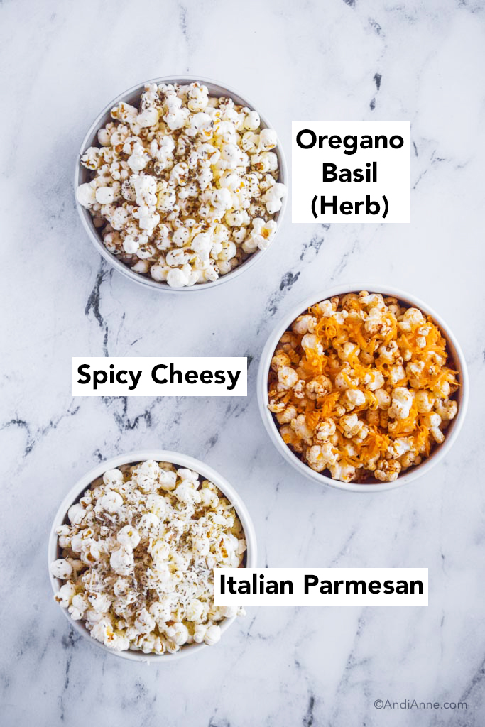 three popcorn seasoning recipes on a counter: bowl of oregano basil herb, spicy cheesy, and italian parmesan popcorn.