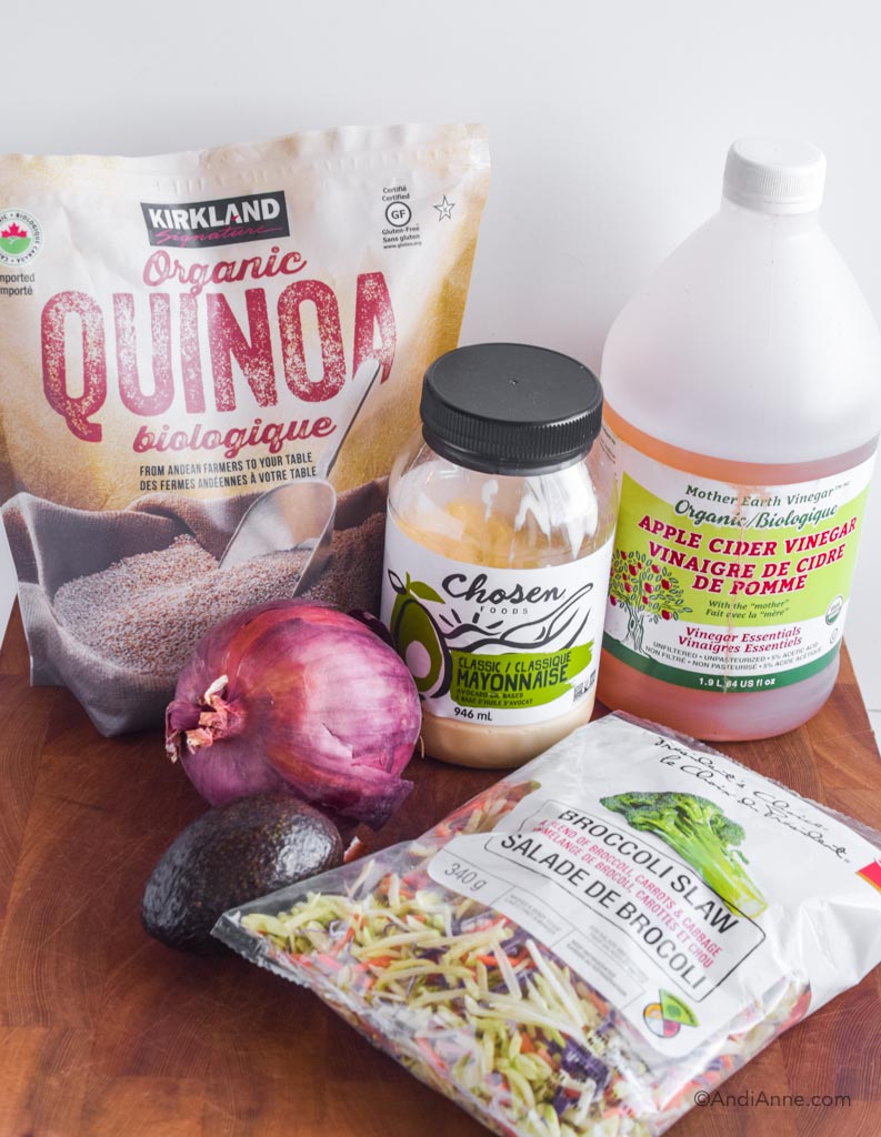 Bag of quinoa, jar of mayonnaise, jug of apple cider vinegar, onion, avocado and bag of broccoli slaw on wood counter.