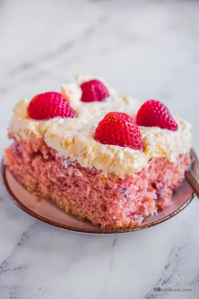 Slice of strawberry pineapple cake on small plate: pink cake, white whipped topping and slices of fresh strawberries on top