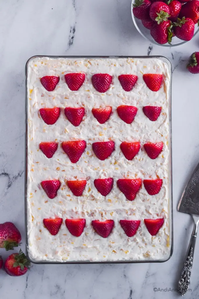 9x13 glass dish with whipped pineapple topping and sliced strawberries in rows. Bowl of fresh strawberries and pie server surround the cake.