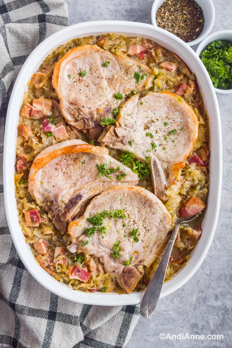Four pork chops overtop of sauerkraut and bacon in a white casserole dish. Small bowls of herbs, kitchen towel and a spoon surround the dish. 