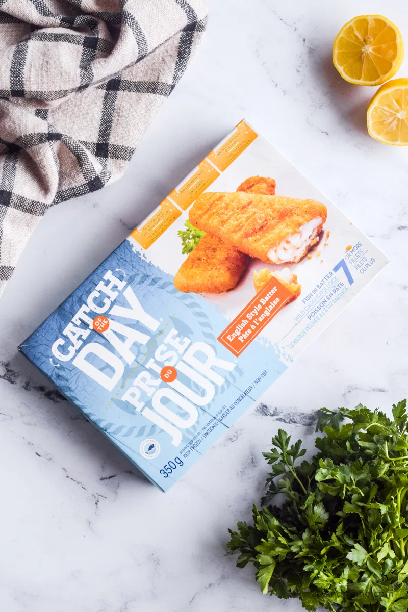 Box of store-bought frozen fish sticks with kitchen towel, fresh parsley and sliced lemon surrounding it. 