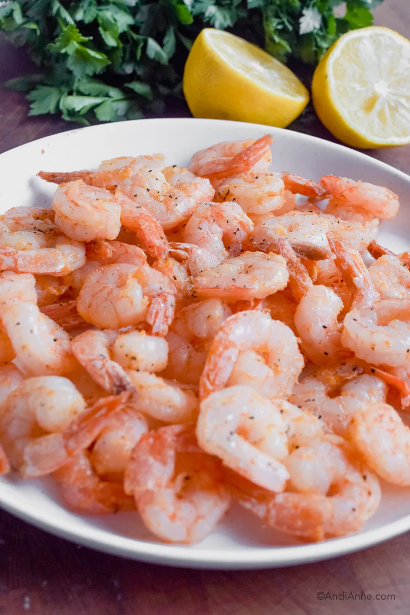 Close up plate of cooked shrimp with sliced lemon and parsley in background.