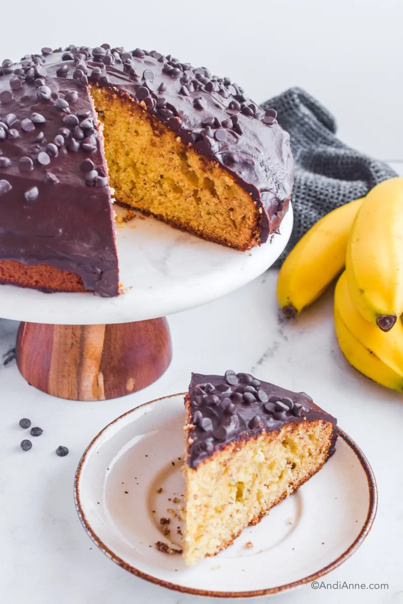 Banana cake with chocolate frosting on cake stand with slice cut out. Plate with slice below it. A few chocolate chips, bananas and a kitchen towel sprinkled around it. 