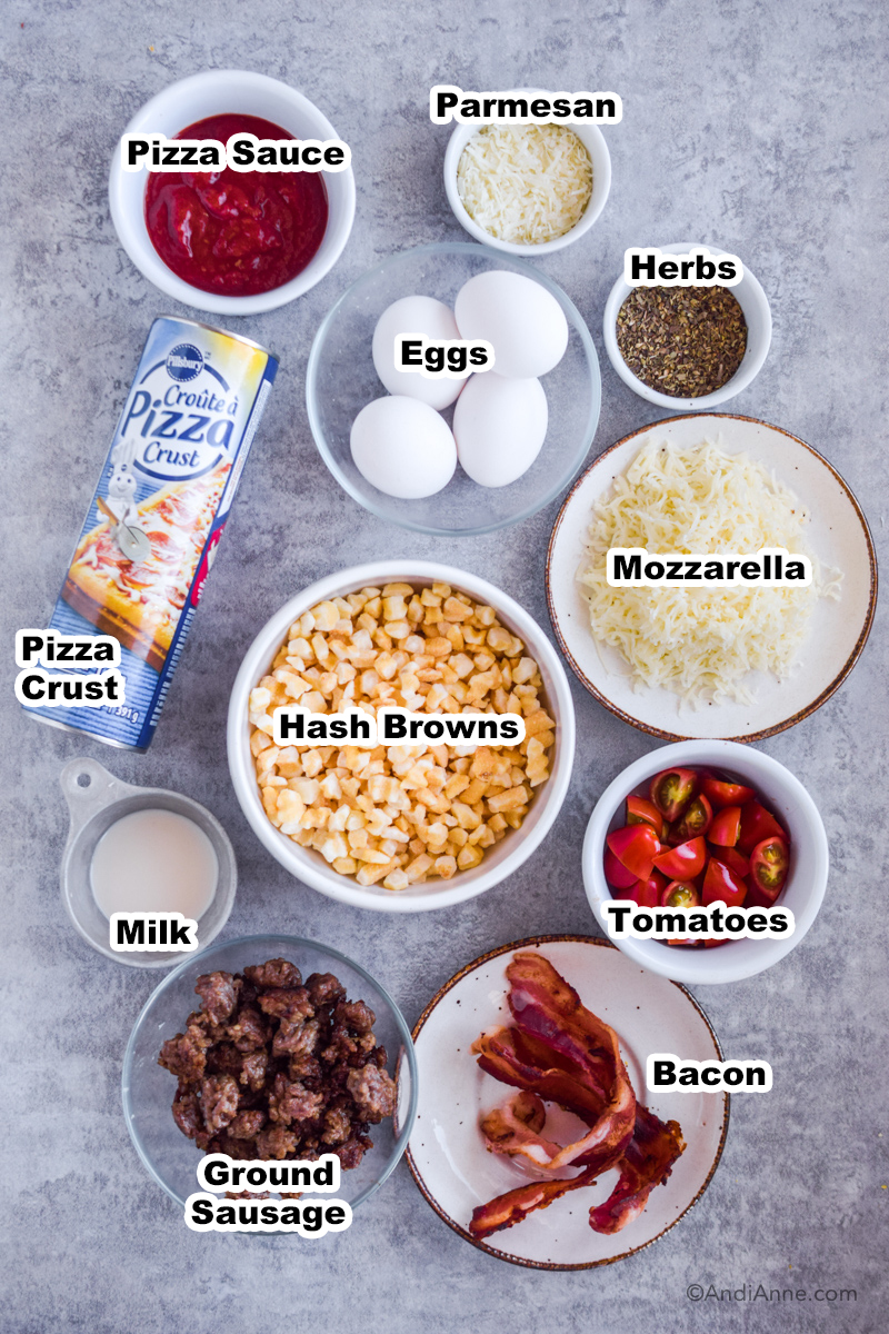 Ingredients used to make breakfast pizza in bowls on the counter including hash browns, pizza crust tube, eggs, bacon and ground sausage meat.