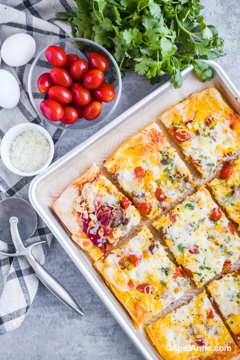 Bacon sausage breakfast pizza on a baking sheet with bowl of grape tomatoes, parsley and cheese beside it.