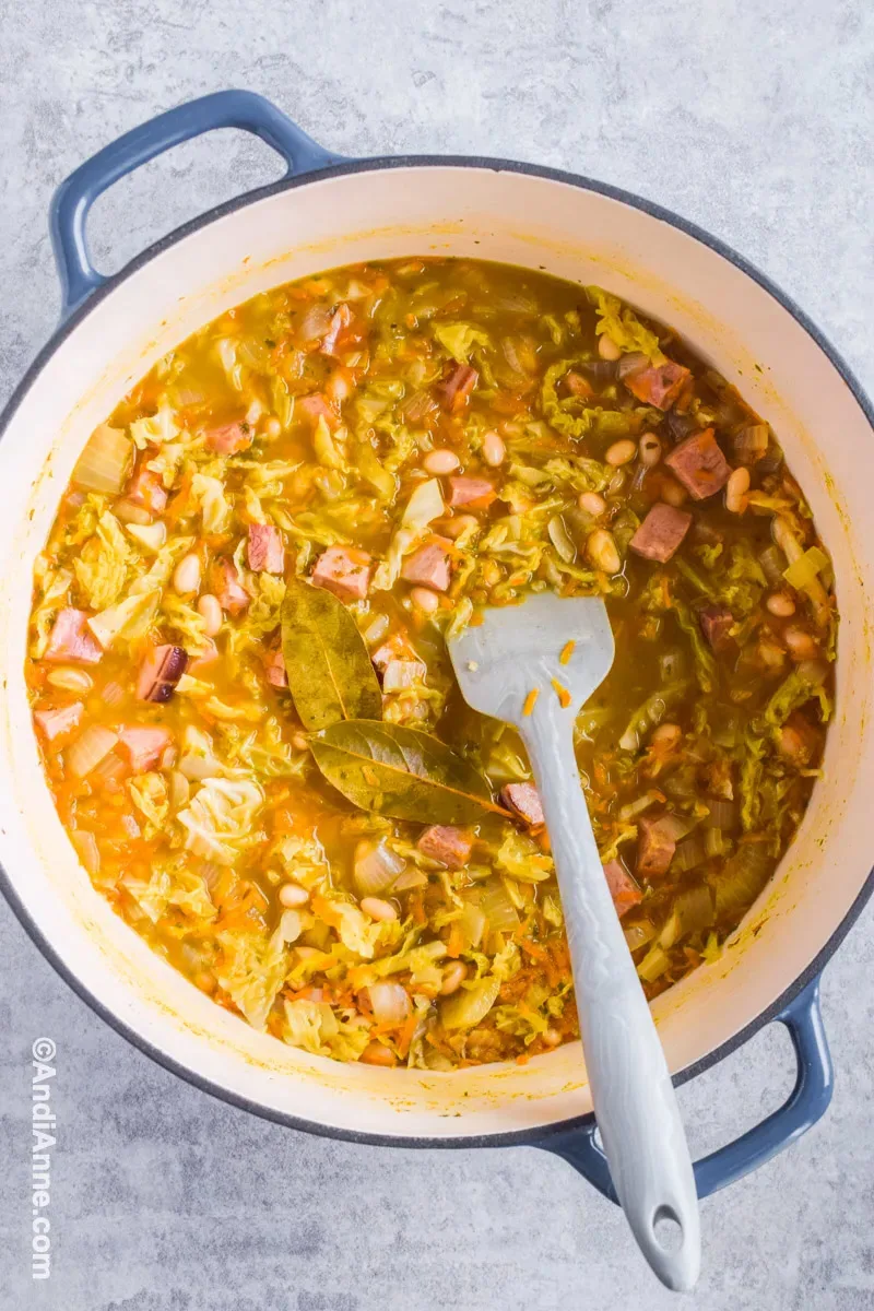 Cooked cabbage ham soup in a large pot with bay leaves and a grey spatula