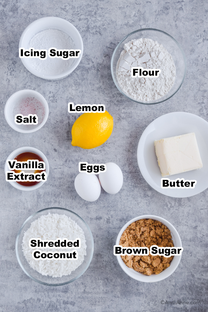 Recipe ingredients on counter including bowl of icing sugar, flour, a lemon, square of butter, two eggs, brown sugar and bowl of shredded coconut.