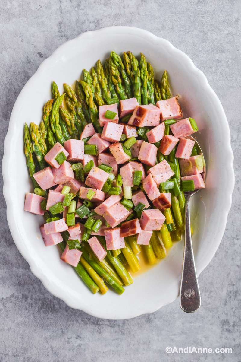 White scalloped bowl with bed of asparagus, chopped ham and green onion on top and a spoon.