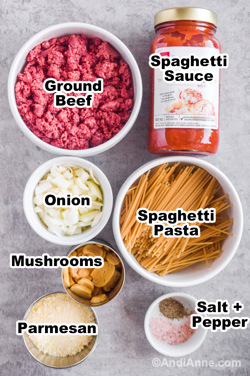 Ingredients for recipe on counter including bowl of raw ground beef, jar of spaghetti sauce, bowl of pasta noodles, chopped onion, canned mushrooms and parmesan.