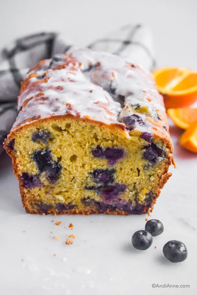 Blueberry orange pound cake with slice cut out so you can see inside. A couple blueberries, sliced orange and kitchen towel surround it.