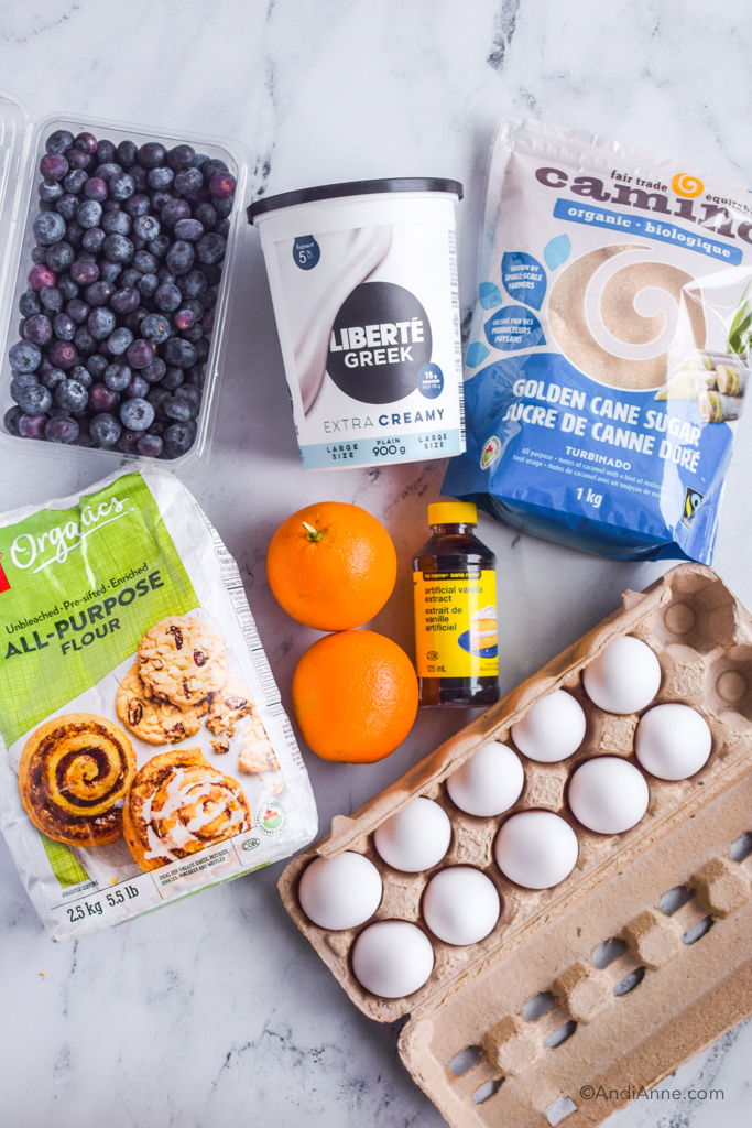 Ingredients lying on counter: carton of fresh blueberries, container of plain yogurt, bag of cane sugar, vanilla extract, two oranges, bag of flour, and carton of eggs.