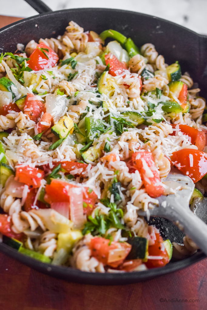 Close up of grated cheese, fresh vegetables and pasta in a skillet.