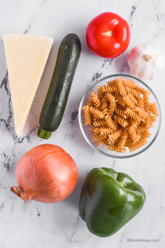 Looking down at ingredients on counter including parmesan cheese, zucchini, tomato, spiral pasta noodles in bowl, garlic bulb, yellow onion, green bell pepper.