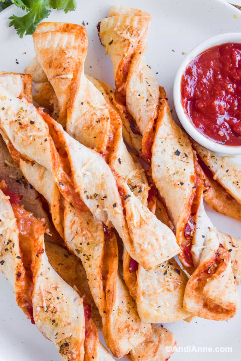 Twisted pizza breadsticks stacked on top of eachother on a white plate with a bowl of pizza sauce.