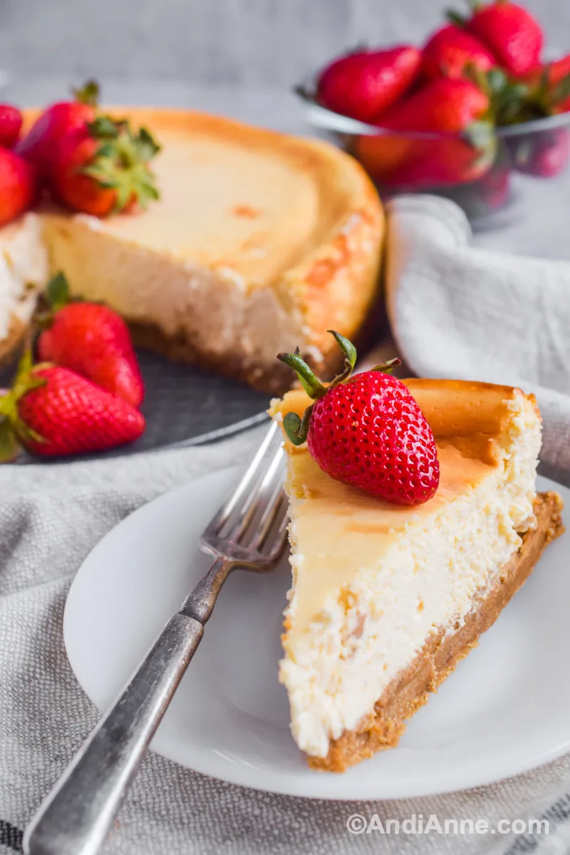 A slice of cheesecake on a plate with a fork and fresh strawberry on top. Full cheesecake and more strawberries piled in the background. 