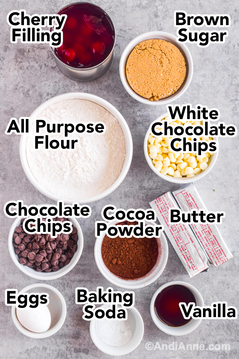 Ingredients to make the recipe on the counter including bowl of flour, brown sugar, cherry pie filling, white chocolate chips, sticks of butter, cocoa powder and vanilla.