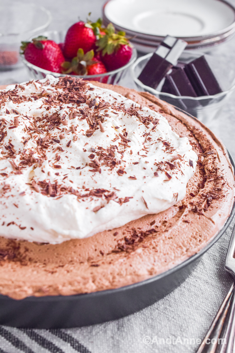 Close up of mocha pie with whipped cream topping and chocolate shavings sprinkled on top. Fresh strawberries and bowl of baker's chocolate squares are blurred in background.