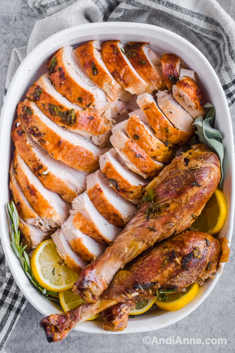 Sliced turkey and drumsticks on a white dish with sliced lemon and fresh rosemary.