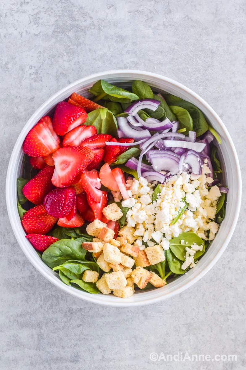 Looking down at large white bowl with strawberries, spinach, red onion, feta, and croutons.