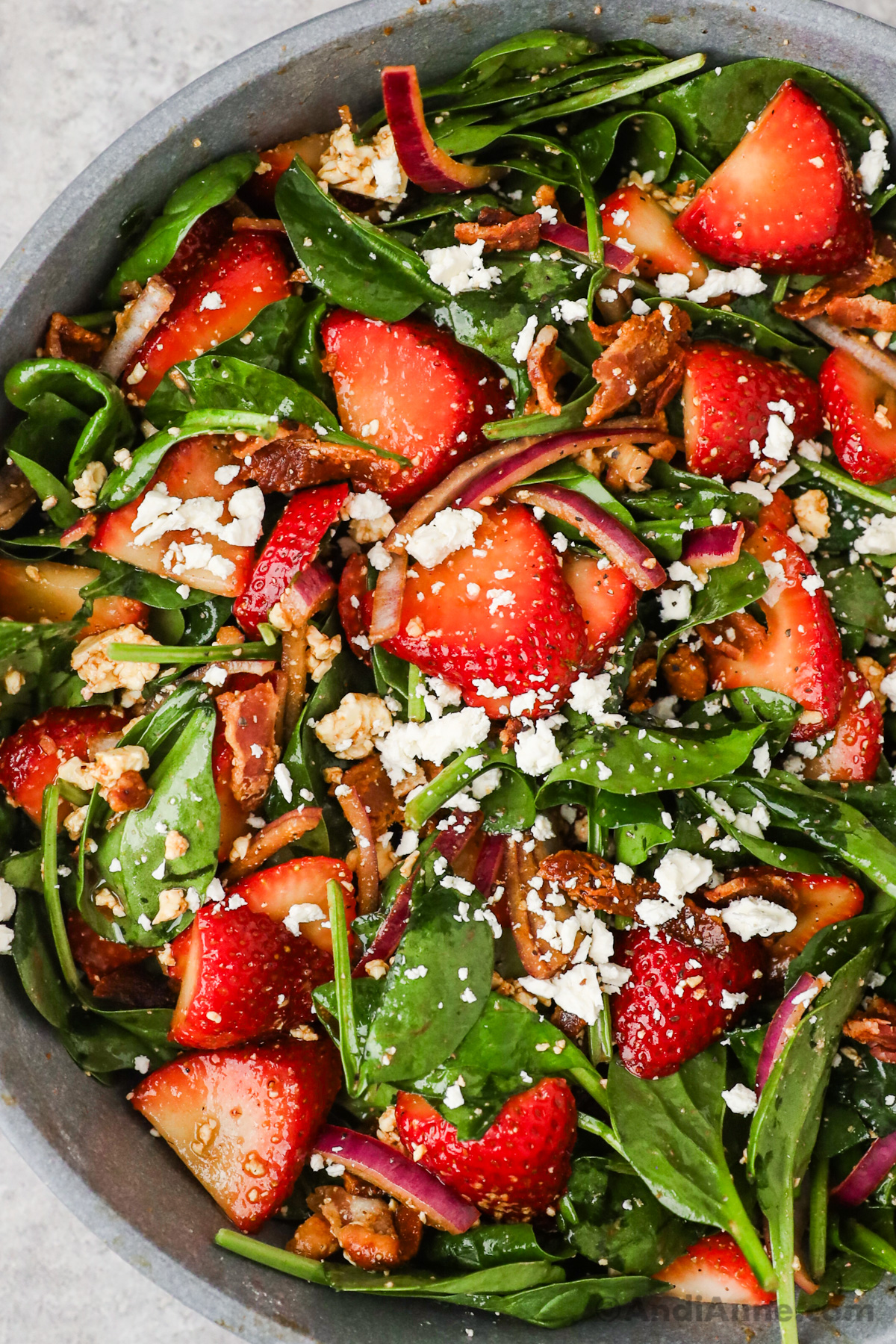 A bowl of strawberry spinach salad with crumbled feta, red onion slices and bacon pieces.