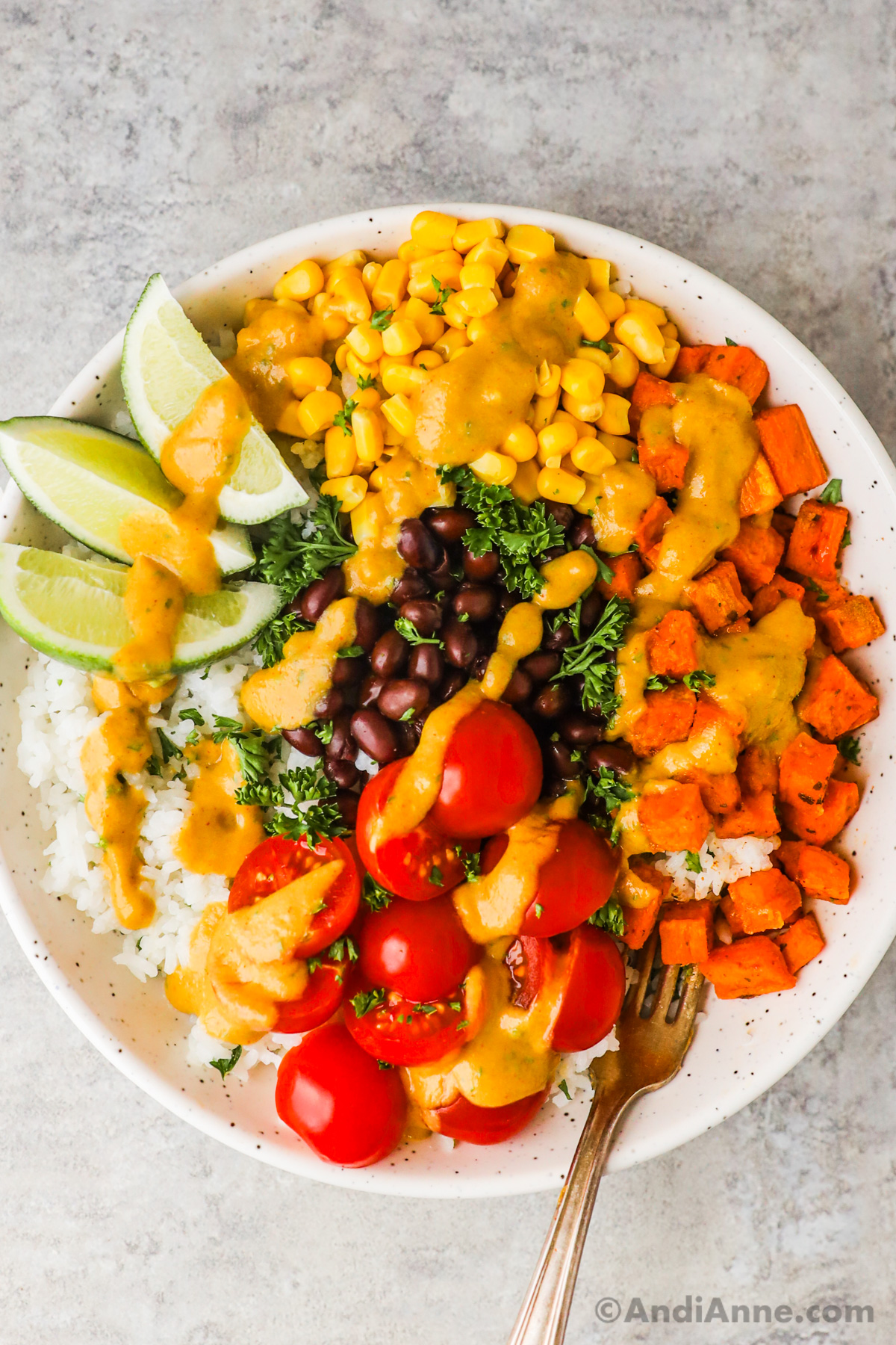 A plate with cubed sweet potatoes, black beans, corn, cherry tomatoes, lime wedges all on a bed of rice, drizzled with creamy avocado dressing.