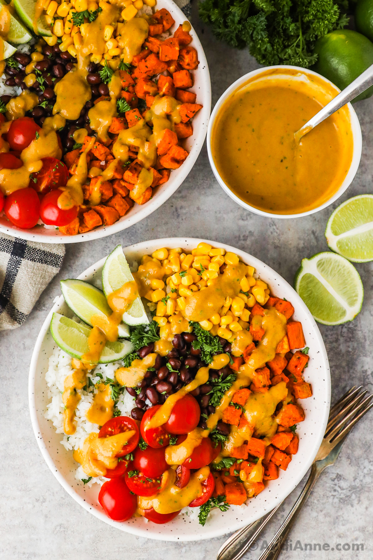 Two plates of sweet potato taco bowl recipe with rice, black beans, corn, tomatoes and lime wedges. All drizzled with a salad dressing.