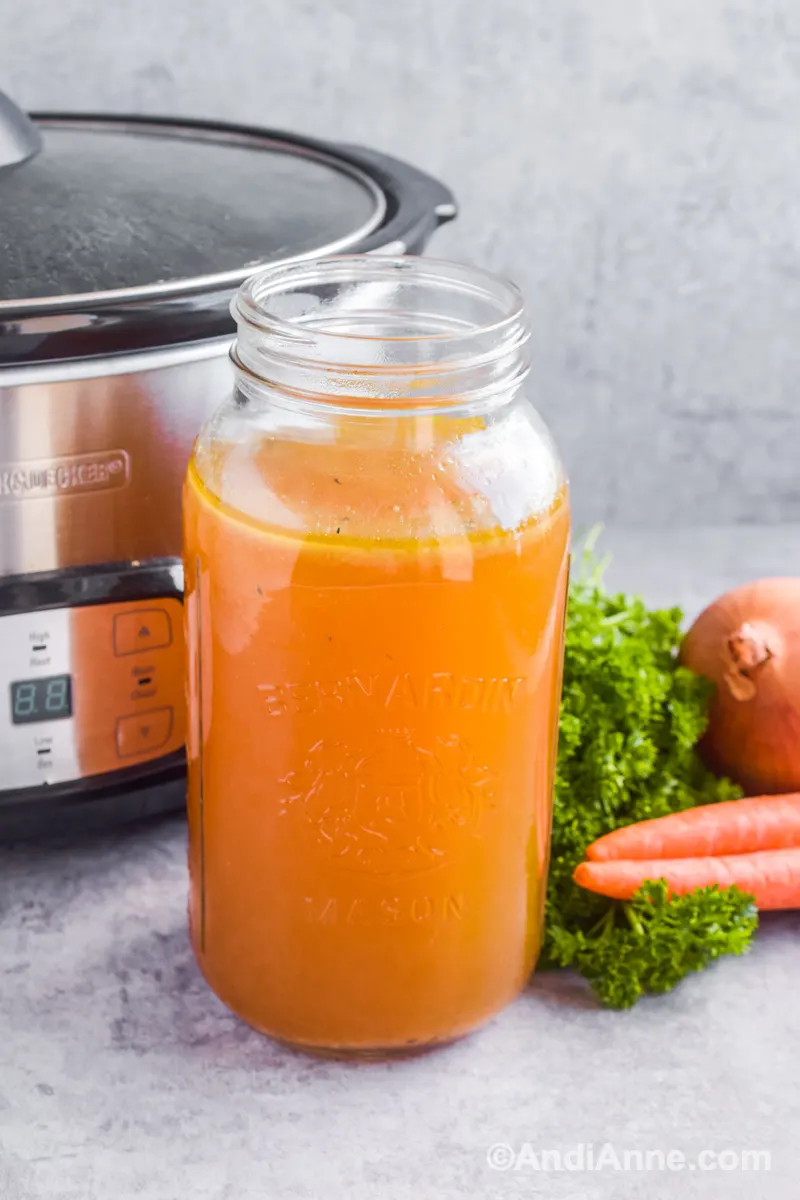 A large mason jar with broth and slow cooker in background.