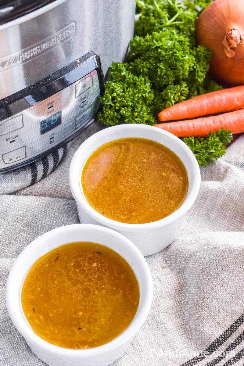 Two small bowls of broth with slow cooker, fresh parsley, carrots and onion in background.