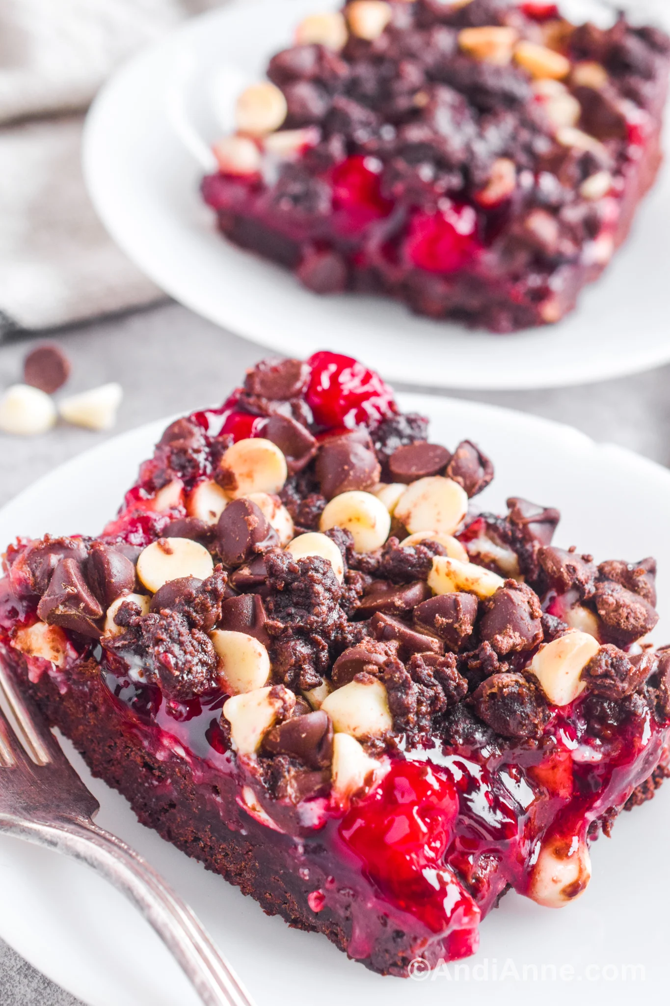 A slice of chocolate cookie cherry bar on a white plate with a fork. Another slice is blurred in the background.
