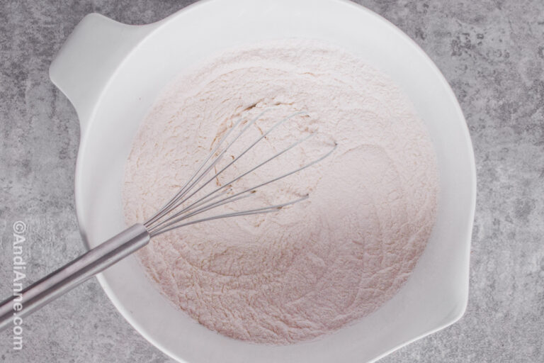 A large white bowl with dry white ingredients including flour and a whisk inside.