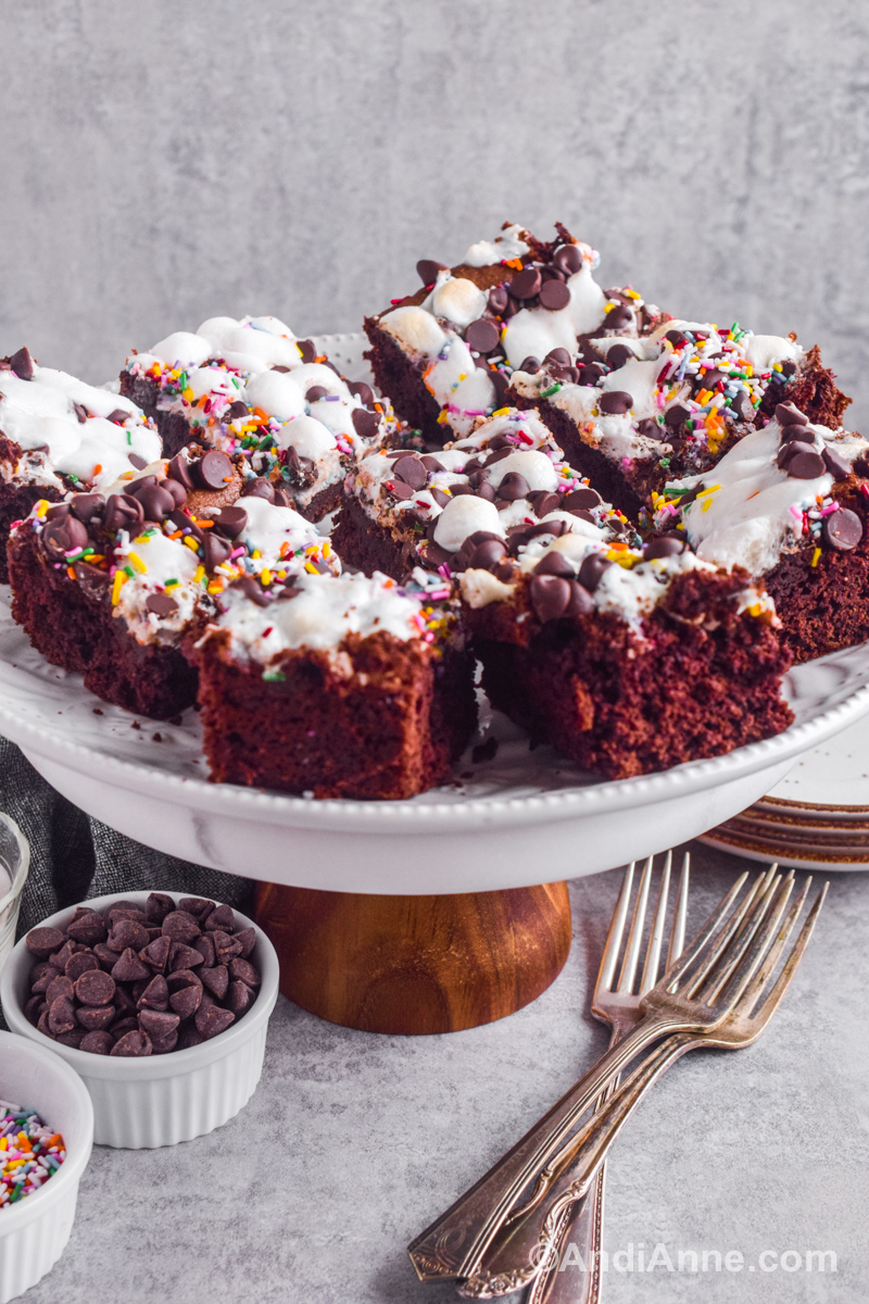 Marshmallow sprinkle cake slices on a serving dish with small bowls of chocolate chips, forks and plates beside it.