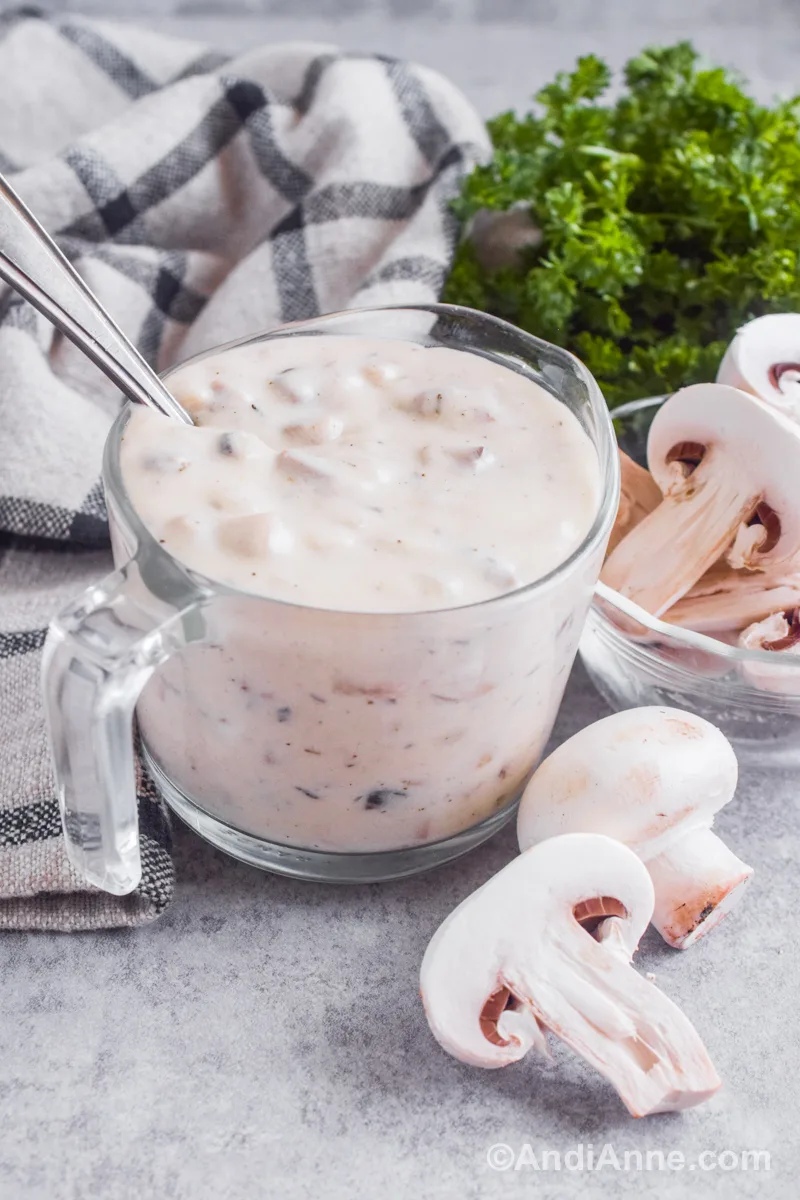 A measuring cup full of white mushroom gravy sauce with a spoon inside. Fresh mushrooms, a kitchen towel and fresh parsley surround the cup.