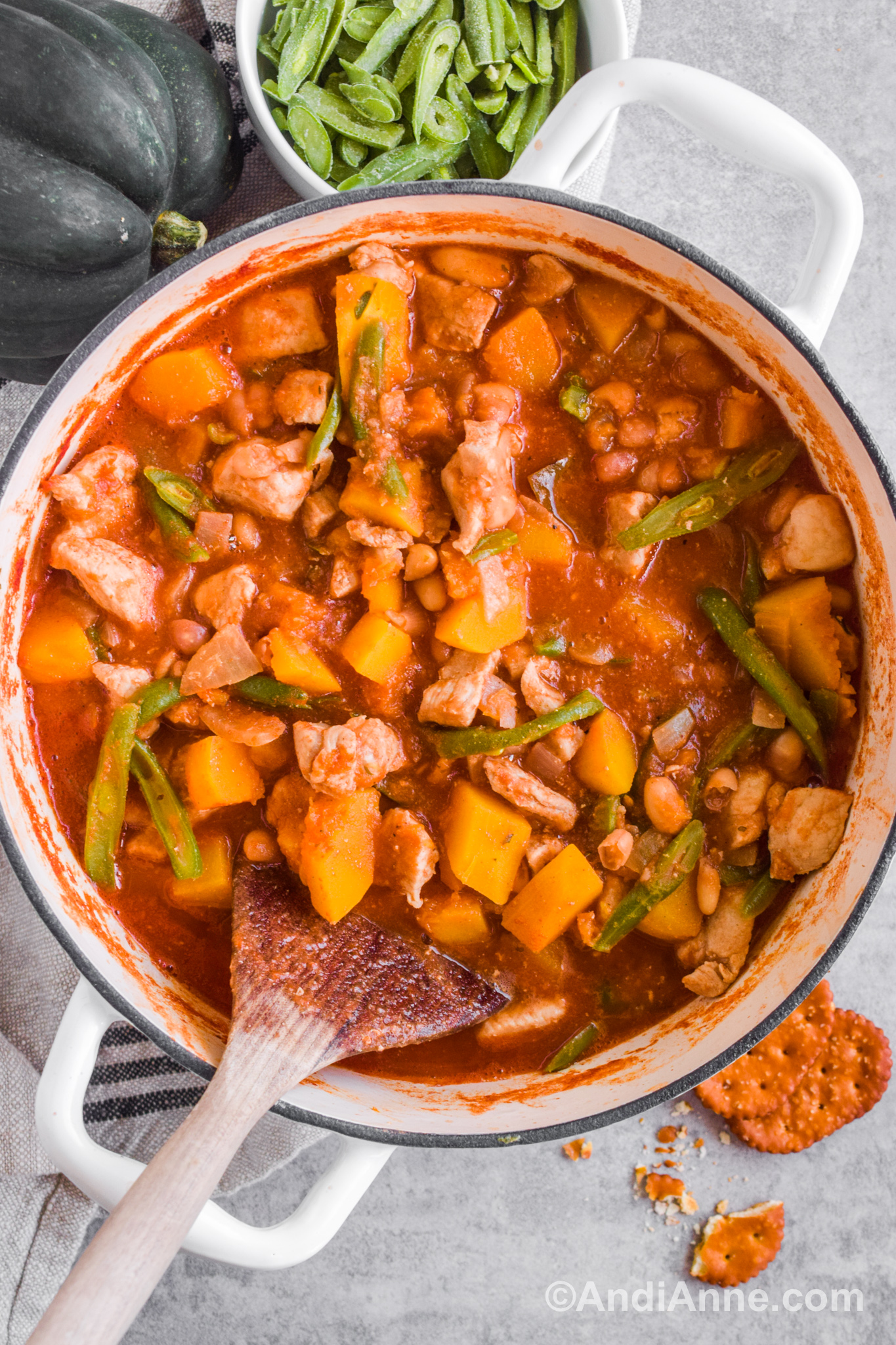 Pork and squash stew in a white pot with a wood spatula. A bowl of green beans, an acorn squash and some crackers surround the bowl.