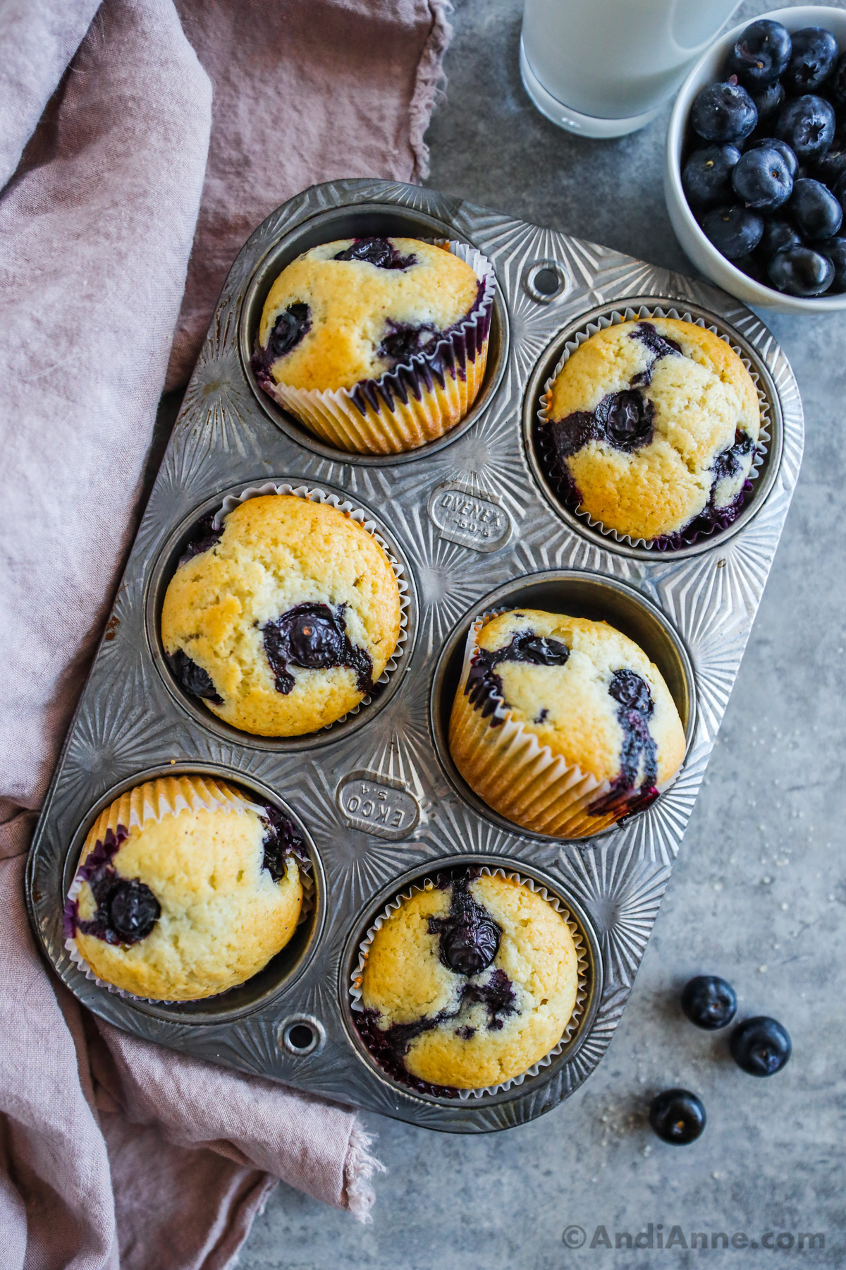 A pan with baked blueberry muffins, a kitchen towel and a bowl of blueberries beside.