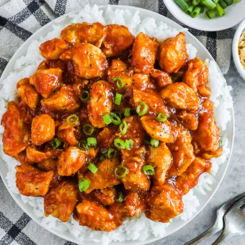 A plated of sweet and sour chicken bites over a bed of rice and chopped green onion on top