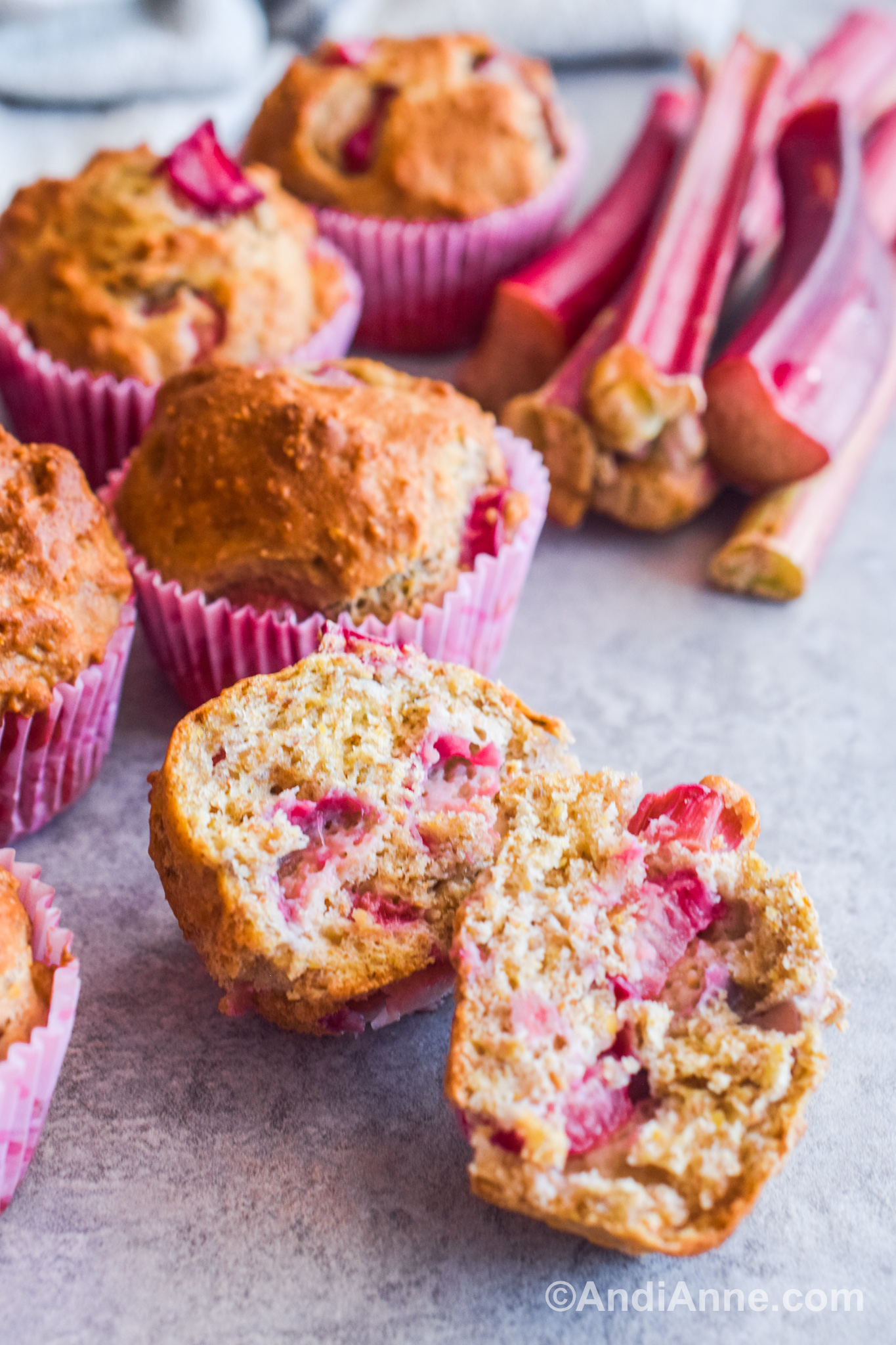 One rhubarb muffin sliced in half with more muffins and rhubarb sticks in background.