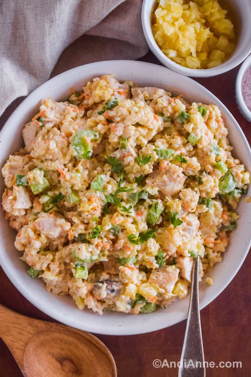 Chicken pineapple and rice salad with chopped parsley in a white bowl with a spoon. Bowl of crushed pineapple and wood spoon surround the bowl.