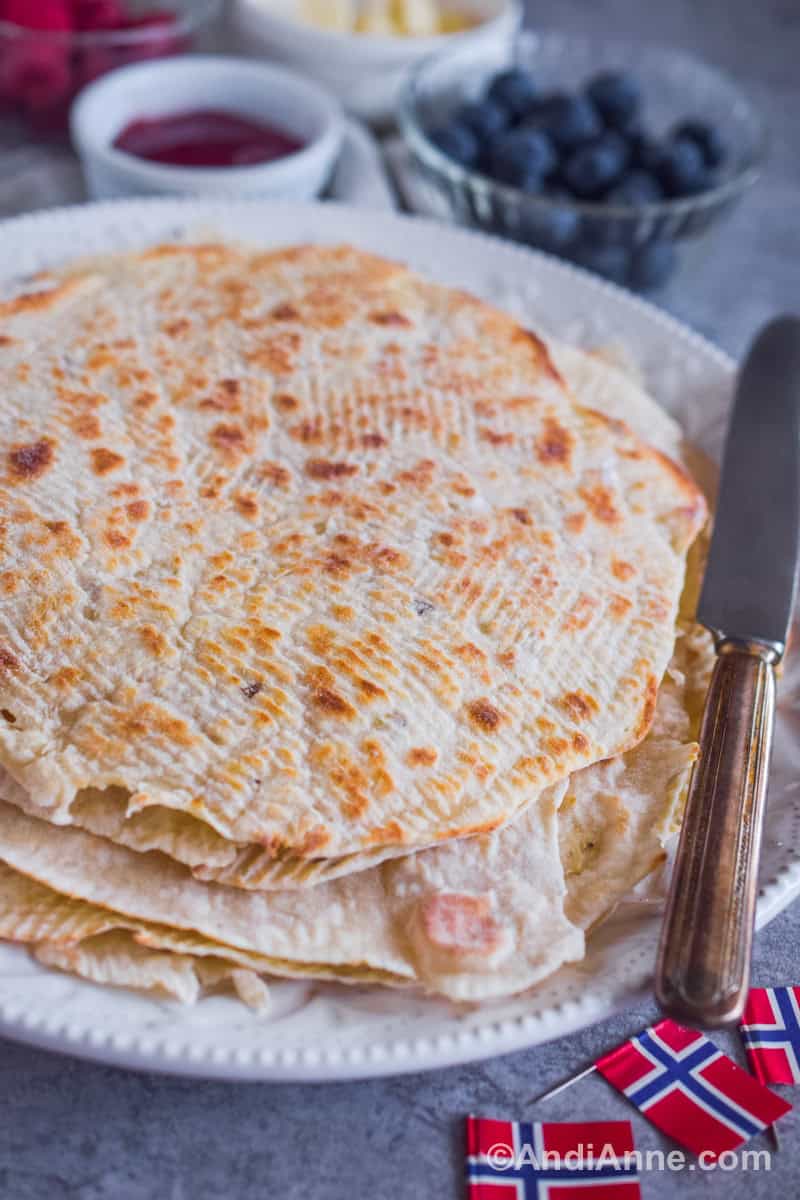 A pile of lefse with a knife, bowls of jam, blueberries and raspberries in background.