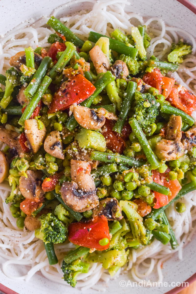 Close up of cooked vegetables including mushrooms, green beans, broccoli, tomatoes, and peas. All sprinkled with grated parmesan and on a bed of spaghetti noodles.