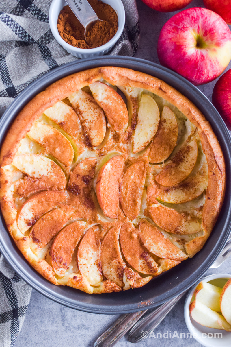 A german apple pancake in a grey cake pan with a small bowl of cinnamon, fresh apples, forks, and a kitchen towel.
