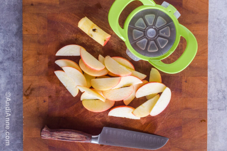 A green apple cutter, sliced apple wedges, an apple core, and a knife on a cutting board.