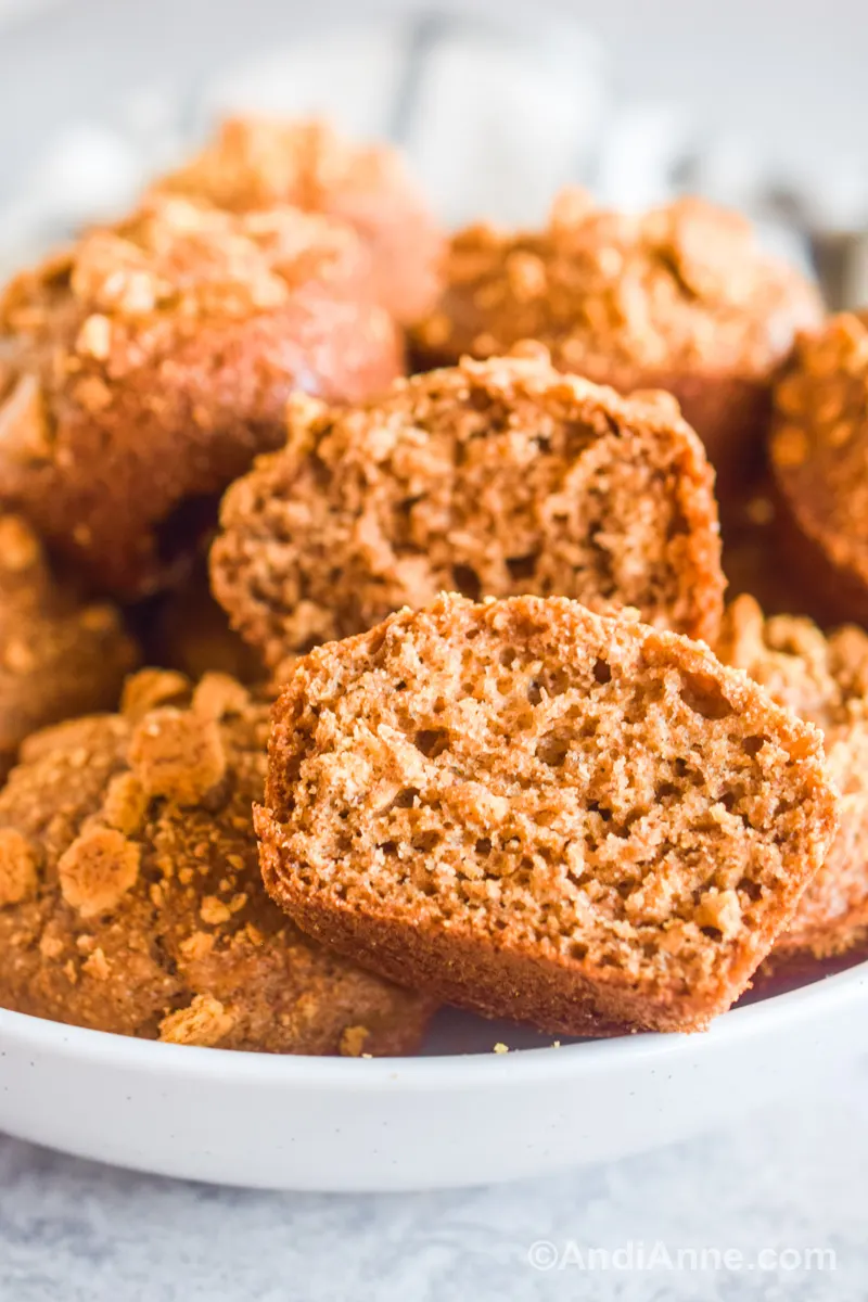 One muffin sliced in half with other muffins stacked behind in a white bowl.