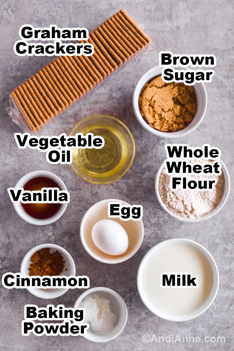 Recipe ingredients on a counter including a bag of graham crackers, and bowls of brown sugar, vegetable oil, vanilla extract, whole wheat flour, milk, baking powder, cinnamon and an egg.