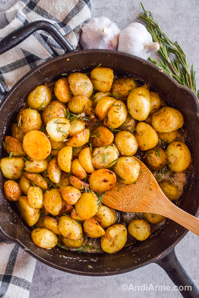 A black skillet with crispy baby potatoes, bubbling oil and butter, and chopped rosemary with a wood spatula.