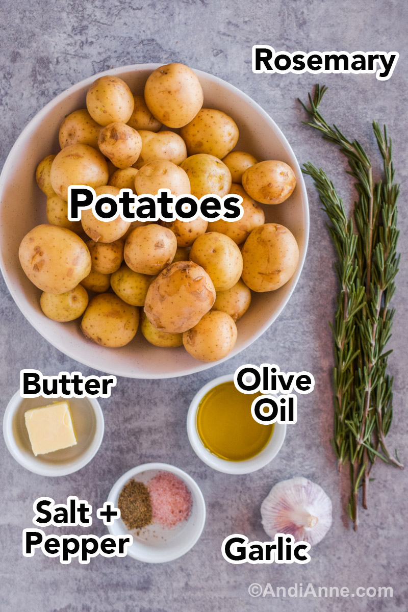 Recipe ingredients on a counter including a bowl of baby potatoes, fresh rosemary sprigs, a garlic bulb, and small bowls with olive oil, butter, salt and pepper.